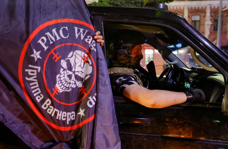 An image of a private mercenary sits in a car next to a Wagner mercenary group flag.