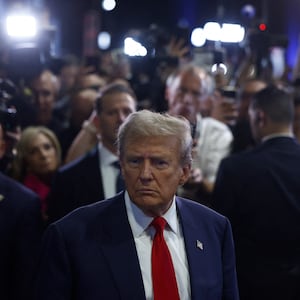 Donald Trump stands withs his back to a busy crowd of people in suits and cameramen, frowning as he looks off into the distance.