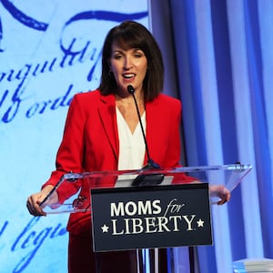 Tina Descovich, wearing red, speaks on stage behind a podium that has a “Moms For Liberty” sign.