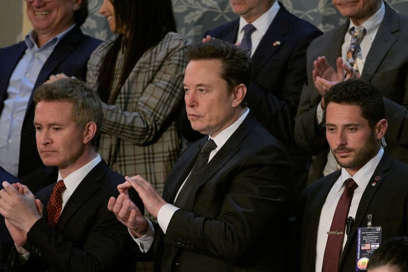 Elon Musk clapping surrounded by other men in suits also clapping