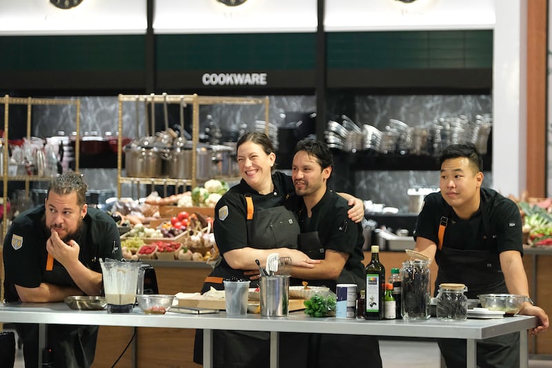 The cast of Top Chef World All Stars stand behind a kitchen island.