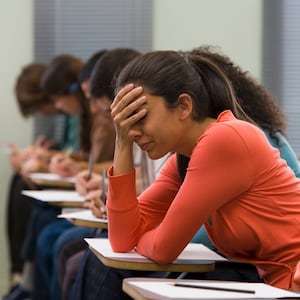 University student wearing a salmon colored top and pony tail sits at desk while taking test with head in her hand. 