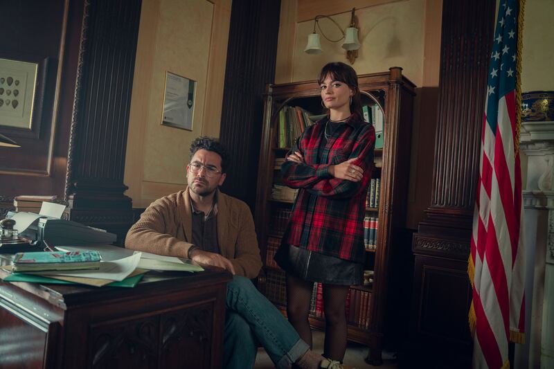 Dan Levy sitting in a chair behind a desk with Emma. Mackey behind him