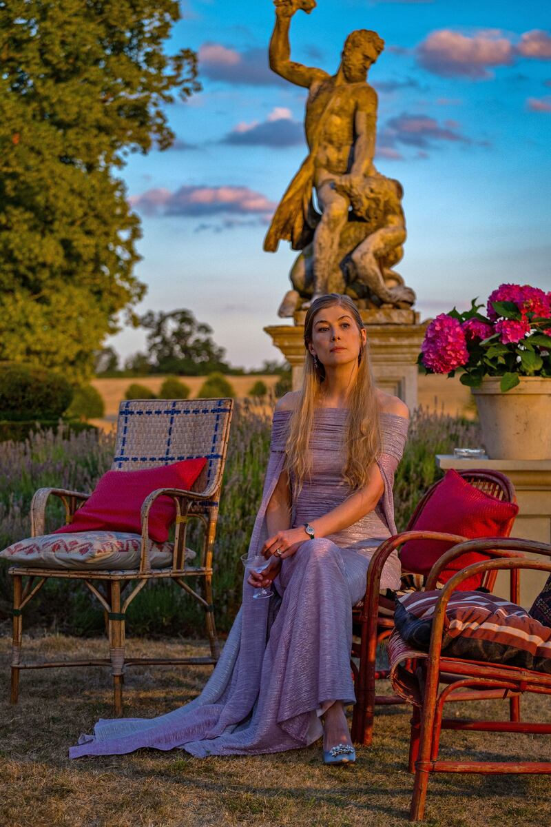 A vertical picture of Rosamund Pike sitting in a chair outside in a still from ‘Saltburn’