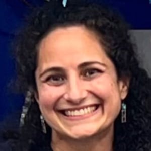 Samantha Woll, wearing a T-shirt and earrings, staring forward smiling. 