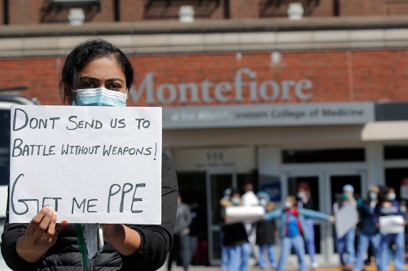 A woman in a mask in front of a hospital holding a sign: Don’t send us to battle without weapons! Get me PPE.