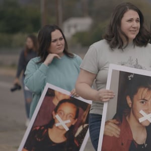 Women march holding pictures of their younger selves with x's crossed over their mouths in a still from 'Let Us Prey'