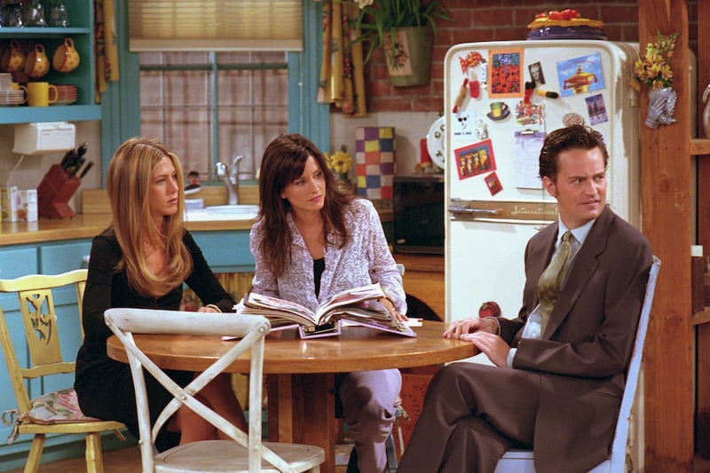 Jennifer Aniston, Courteney Cox and Matthew Perry sit together at a table on the set of "Friends".