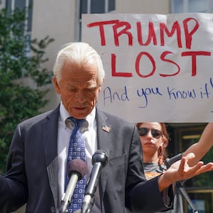 Peter Navarro, wearing a suit, stares down during a press conference where he was heckled by a protester holding a “TRUMP LOST” sign.