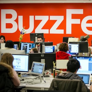 BuzzFeed employees work at their desks behind a BuzzFeed logo in red.