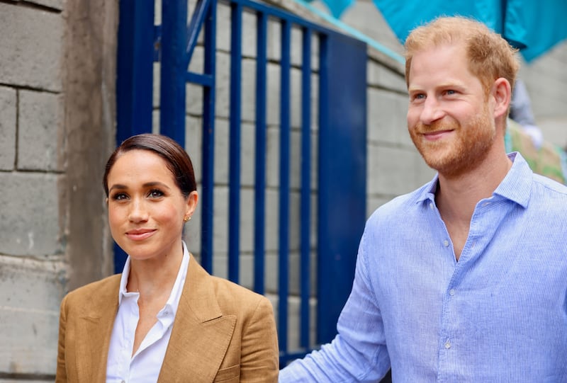 Prince Harry in a blue linen short smiling beside Meghan Markle