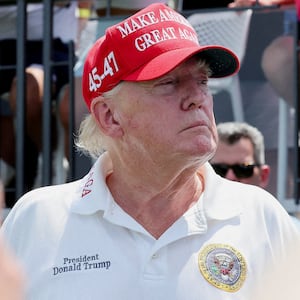 Donald Trump at a golf course wearing a red MAGA hat and a white polo shirt.