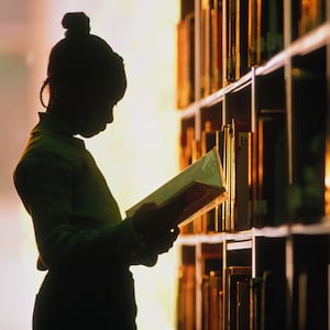 Young school girl stands holding a library book silhouetted next to a library book shelf. 