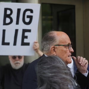 Rudy Giuliani, wearing a suit, leaves a courthouse flanked by protesters, including one holding a sign that says, “BIG LIE.”