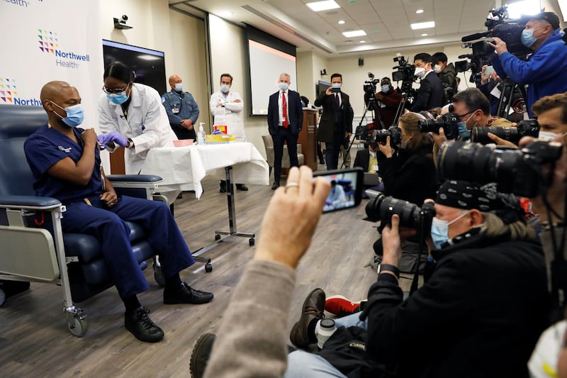 A photo of Dr. Yves Duroseau receiving one of the first COVID vaccines, as a throng of news photographers document the occasion.