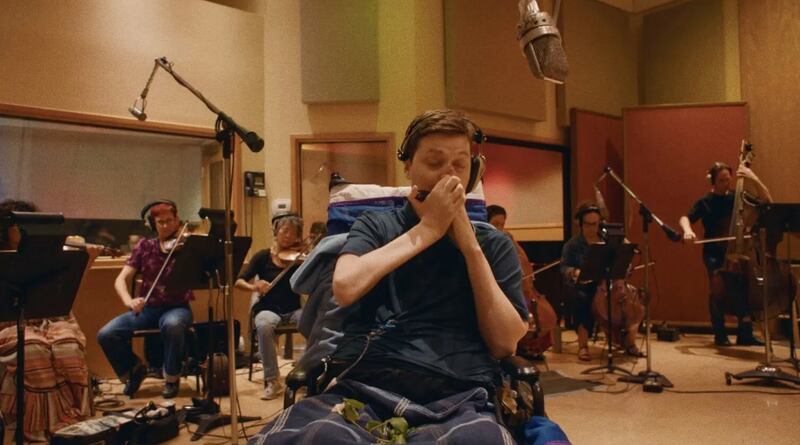 Mac Sinise plays his harmonica as he sits in a wheelchair during a set.