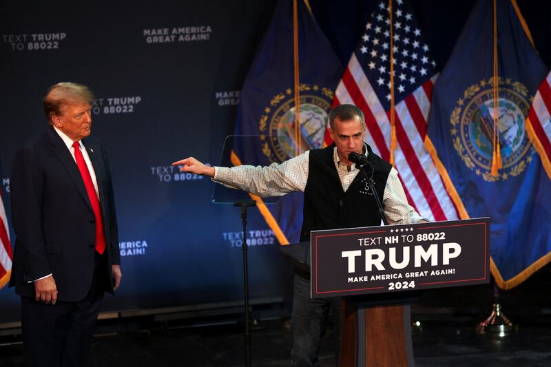 Corey Lewandowski speaks behind a podium, next to Donald Trump