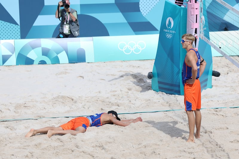 A beach volleyball player in orange top and blue shorts lies on the sand while to his right a similarly dressed player stands 