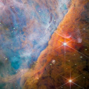  A nebula made of many layers of cloudy, colourful material. The top-left side of the image is brightly lit, filled with wispy, thin material in pale shades of pink and blue. A thick bar of denser, cloudier material crosses diagonally at the bottom right.